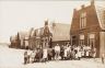 1e Woudstraat. Bewoners poseren omstreeks 1915 trots in hun nieuwe straat. recht vooraan het pand van Kapper Lenters, knippen 15 cent, zelf bloempot meenemen. Na de ruimte het woonhuis met de wasserij van Hoeksema. Dacostaschool en Chroom en nikkelfabriek Batstra en Fritsma.