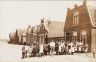 1e Woudstraat. Foto van omstreeks 1914, er werd nog aan de bestrating gewerkt. Achter de links staande kinderen, met bord op de muur, de boordenwasserij en strijkinrichting van Hoeksema. Het koepeltje daarnaast was de ingang van de 2 klassige 'Da Costa' school. Daarnaast de chroom en emailleer werkplaats van Batstra. 